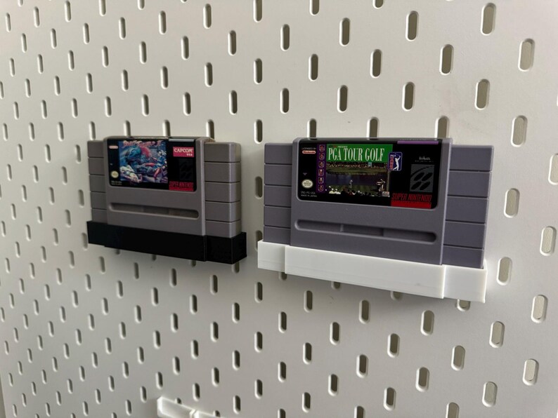 K&ouml;nnte beinhalten: Zwei graue Super Nintendo-Spielmodule. Eines hat einen schwarzen Sockel, das andere einen wei&szlig;en Sockel. Die Spielmodule sind an einer wei&szlig;en Lochwand befestigt. Ein Modul tr&auml;gt den Text "PGA TOUR GOLF".