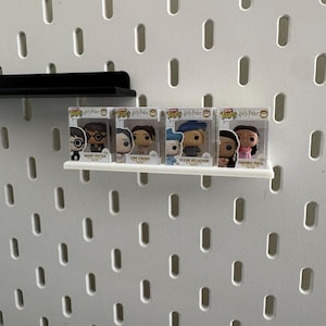 Puede incluir: Una estantería blanca exhibe cuatro figuras Funko Pop! de la serie Harry Potter. Las figuras están en cajas de plástico transparente, y la estantería está montada en una pared de tablero perforado blanco.