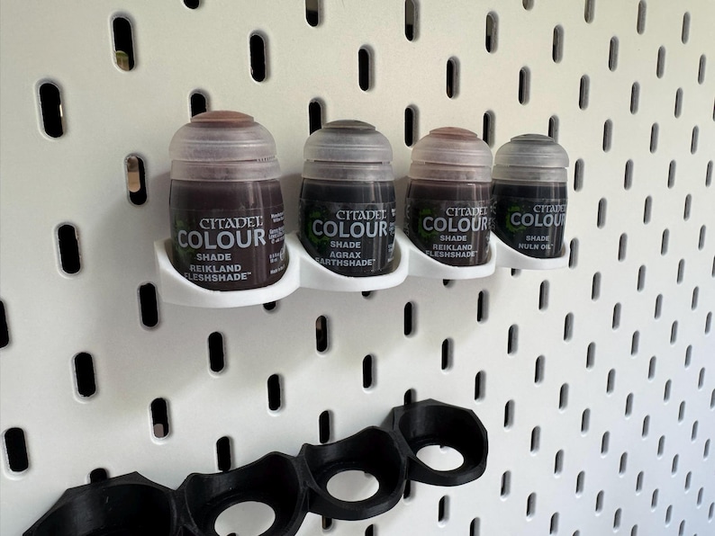 Puede incluir: Cuatro peque&ntilde;os frascos de pintura Citadel Colour, tonos Reikland Fleshshade, Agrax Earthshade, Reikland Fleshshade y Nuln Oil, se muestran en una pared de tablero de clavijas blanco con un soporte de pintura de pl&aacute;stico negro debajo.