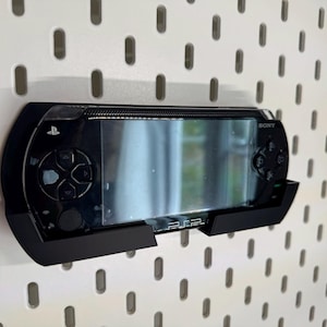 Może przedstawiać: Czarna konsola do gier przenośna Sony PSP zamontowana na czarnym wsporniku na białej ścianie z otworami. PSP jest wyśrodkowane w kadrze, wyświetlając ekran i przyciski sterowania. Wspornik zapewnia wsparcie dla urządzenia.