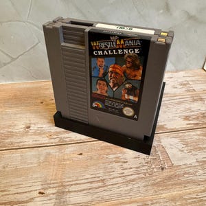 May include: A gray Nintendo Entertainment System (NES) video game cartridge for WWF WrestleMania Challenge. The cartridge features a colorful label with images of wrestlers. The cartridge is in a black stand.