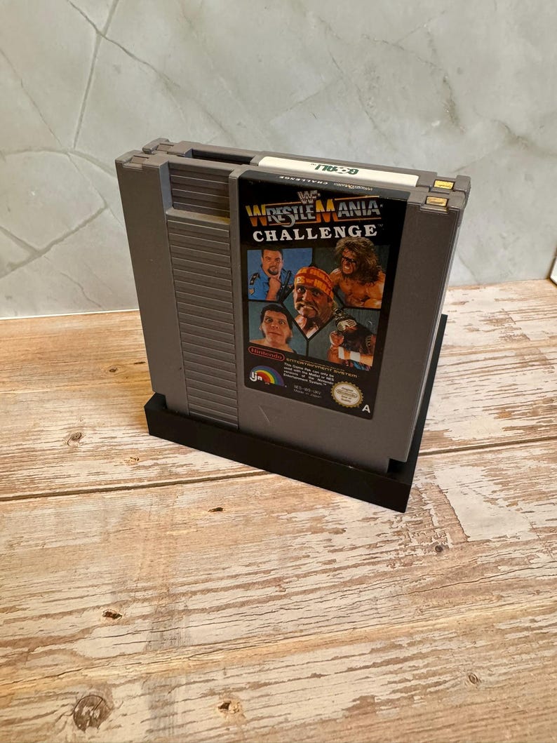 May include: A gray Nintendo Entertainment System (NES) video game cartridge for WWF WrestleMania Challenge. The cartridge features a colorful label with images of wrestlers. The cartridge is in a black stand.