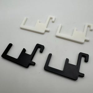Puede incluir: Cuatro objetos de pl&aacute;stico en forma de gancho. Dos son blancos y dos son negros. Cada uno tiene una base rectangular con un poste vertical y un gancho curvo. Los objetos est&aacute;n dispuestos sobre una superficie blanca.