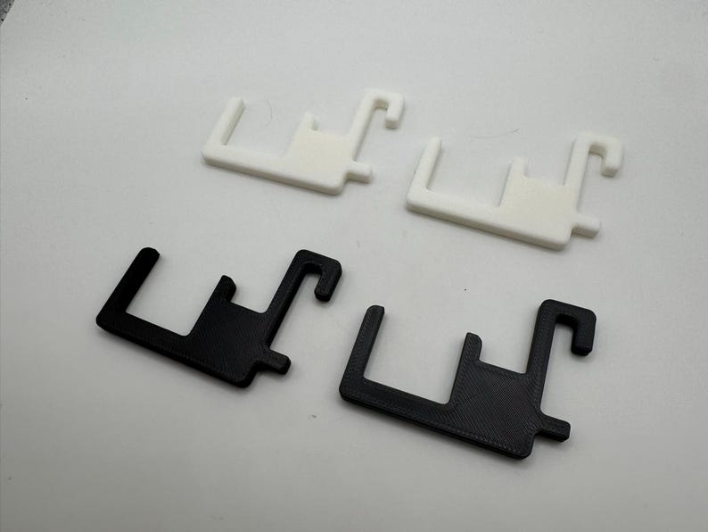 Puede incluir: Cuatro objetos de pl&aacute;stico en forma de gancho. Dos son blancos y dos son negros. Cada uno tiene una base rectangular con un poste vertical y un gancho curvo. Los objetos est&aacute;n dispuestos sobre una superficie blanca.