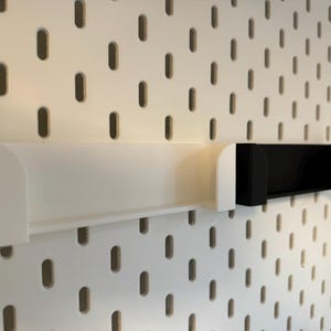 Puede incluir: Estantes blancos y negros para panel perforado. El estante blanco es largo y rectangular, con bordes curvos. El estante negro es m&aacute;s corto y tiene un dise&ntilde;o en forma de U. Ambos estantes est&aacute;n montados en un panel perforado blanco con agujeros ovalados.