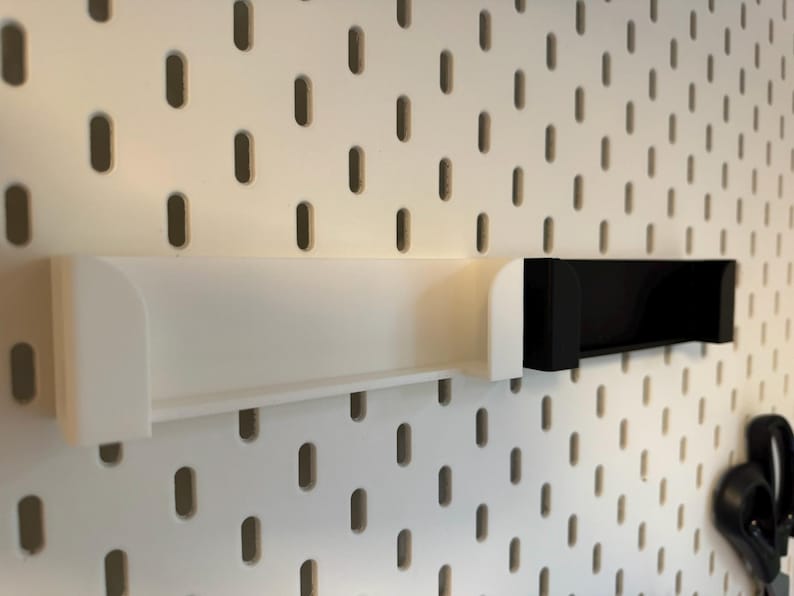 Puede incluir: Estantes blancos y negros para panel perforado. El estante blanco es largo y rectangular, con bordes curvos. El estante negro es m&aacute;s corto y tiene un dise&ntilde;o en forma de U. Ambos estantes est&aacute;n montados en un panel perforado blanco con agujeros ovalados.