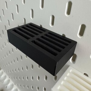 Op de afbeelding: Een zwart rechthoekig schap met een sleufontwerp, gemonteerd op een wit pegboard. Het schap is gemaakt van een solide materiaal en is ontworpen om items vast te houden. Het pegboard heeft gelijkmatig verdeelde gaten.