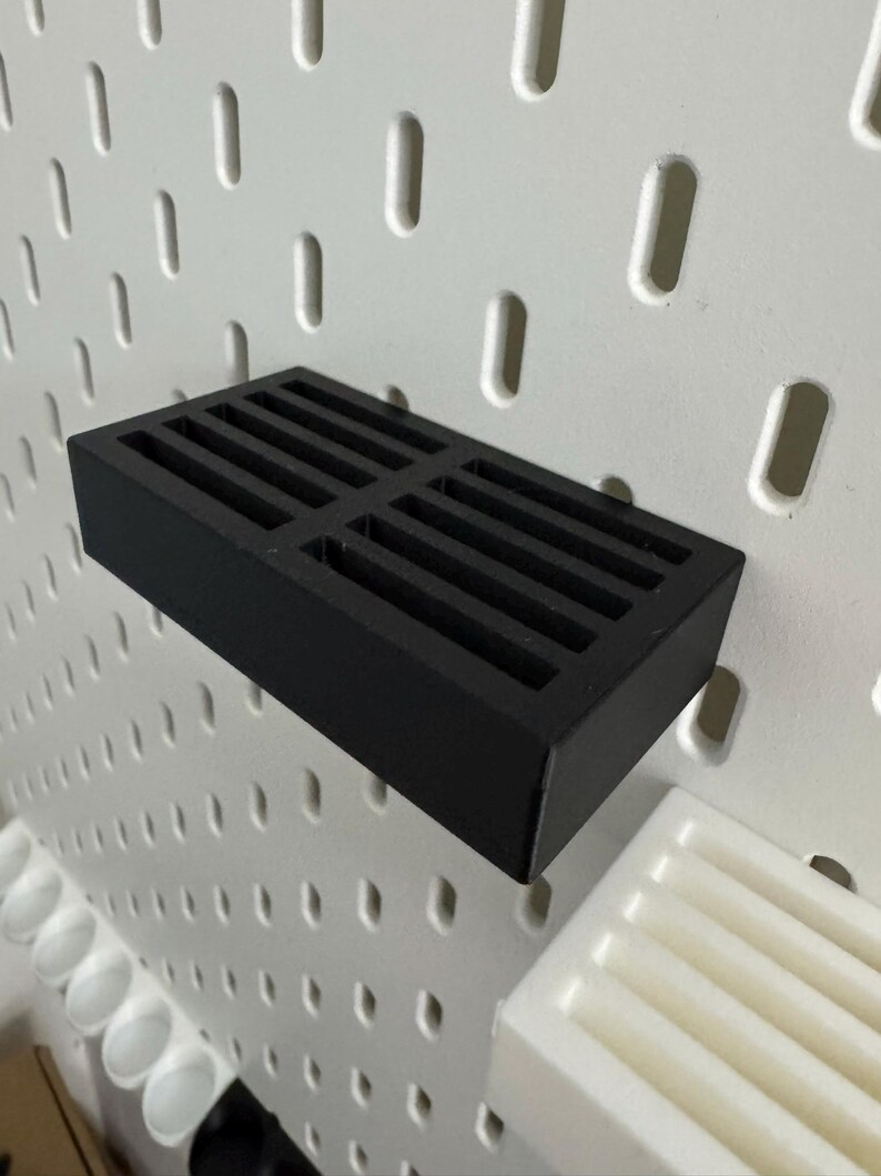 Op de afbeelding: Een zwart rechthoekig schap met een sleufontwerp, gemonteerd op een wit pegboard. Het schap is gemaakt van een solide materiaal en is ontworpen om items vast te houden. Het pegboard heeft gelijkmatig verdeelde gaten.