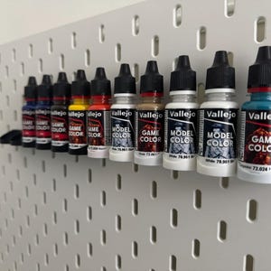 Op de afbeelding: Een verzameling Vallejo-verfpotjes in verschillende kleuren, waaronder rood, geel en wit, wordt tentoongesteld op een wit pegboard. De potjes zijn gelabeld met "Game Color" en "Model Color". De potjes zijn in een rij gerangschikt, met zwarte doppen en witte etiketten.
