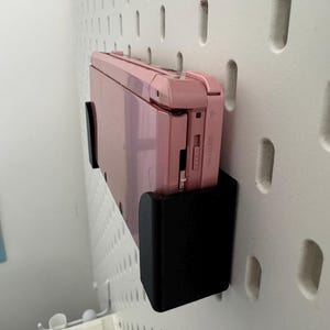 Pode incluir: Um console de jogos port&aacute;til Nintendo 3DS rosa est&aacute; montado em uma parede de placa de pinos branca usando um suporte preto impresso em 3D.