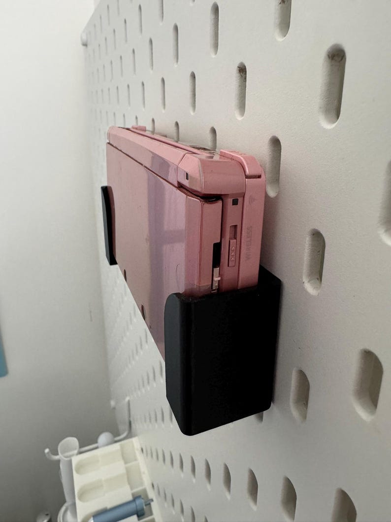 Pode incluir: Um console de jogos port&aacute;til Nintendo 3DS rosa est&aacute; montado em uma parede de placa de pinos branca usando um suporte preto impresso em 3D.