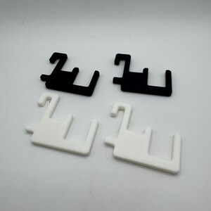May include: Four plastic clips, two black and two white, are arranged on a white surface. Each clip has a unique shape with a hook and two perpendicular arms, suggesting a fastening or hanging function. The clips are evenly spaced and well-lit.