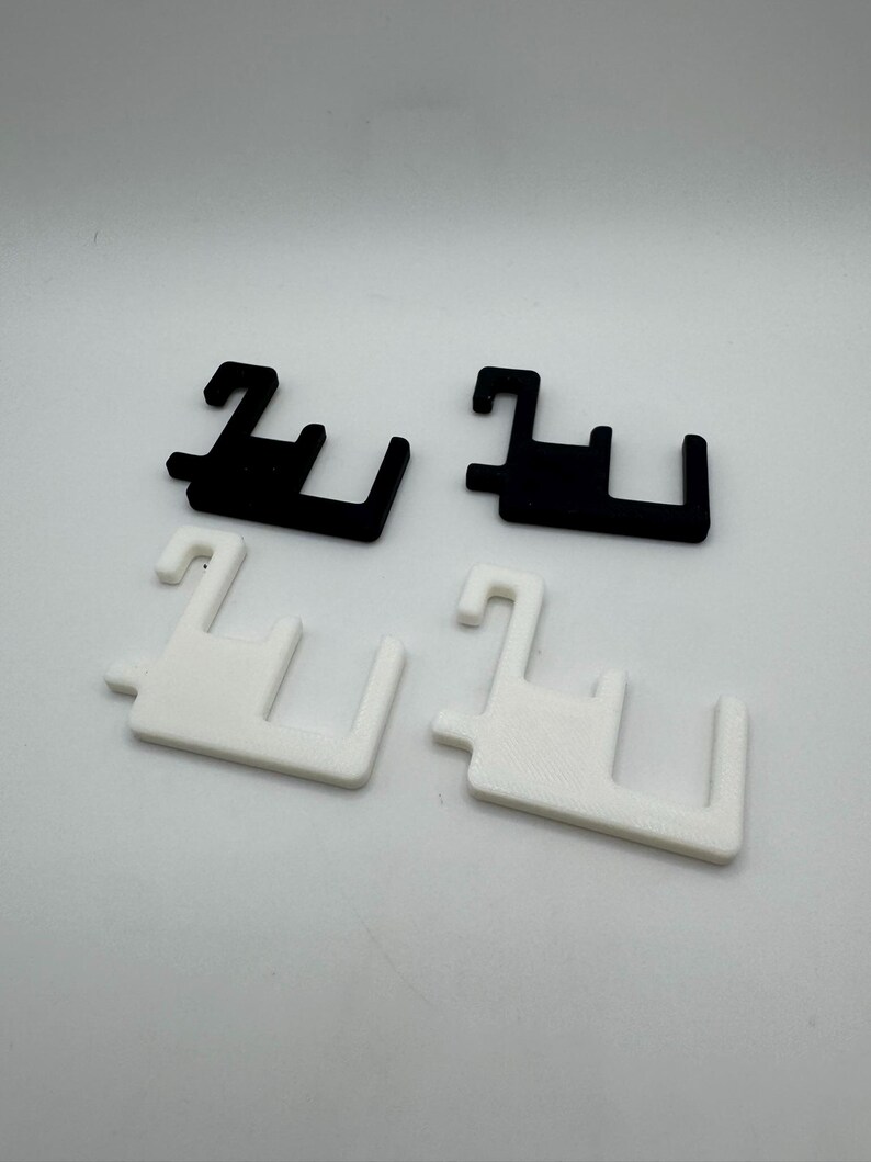 May include: Four plastic clips, two black and two white, are arranged on a white surface. Each clip has a unique shape with a hook and two perpendicular arms, suggesting a fastening or hanging function. The clips are evenly spaced and well-lit.