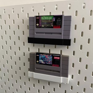 K&ouml;nnte beinhalten: Zwei Super Nintendo-Videospielkassetten, die auf einer wei&szlig;en Lochwand ausgestellt sind. Die obere Kassette ist mit "PGA Tour Golf" mit einem gr&uuml;nen und lila Etikett beschriftet. Die untere Kassette hat ein farbenfrohes Etikett mit dem Capcom-Logo.