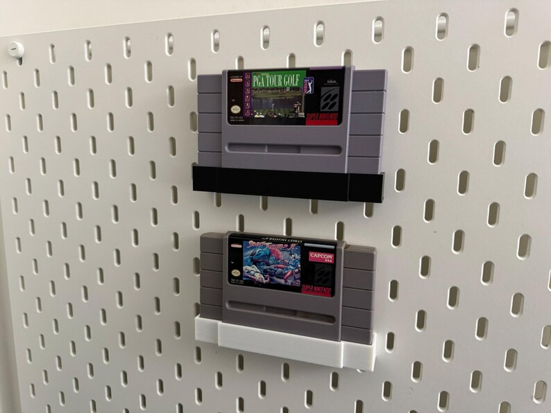 K&ouml;nnte beinhalten: Zwei Super Nintendo-Videospielkassetten, die auf einer wei&szlig;en Lochwand ausgestellt sind. Die obere Kassette ist mit "PGA Tour Golf" mit einem gr&uuml;nen und lila Etikett beschriftet. Die untere Kassette hat ein farbenfrohes Etikett mit dem Capcom-Logo.