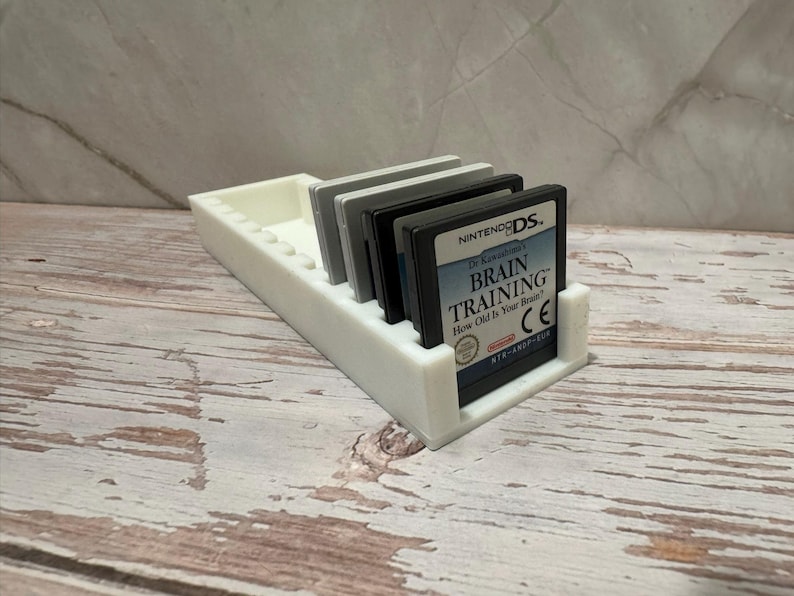 Puede incluir: Un soporte de cartuchos de juego de pl&aacute;stico blanco con varios cartuchos de juego de Nintendo DS. Los cartuchos de juego son negros, grises y blancos. Un cartucho muestra el texto "Brain Training".