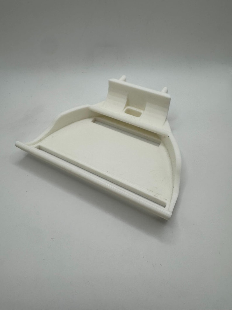May include: A white, 3D-printed scoop-shaped object with a handle and a raised back. The scoop has a curved front and a rectangular opening in the back. The object is made of a solid, opaque material.