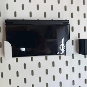 Puede incluir: Una consola de juegos portátil negra está montada en una pared perforada blanca con un soporte blanco. La consola está cerrada y tiene una superficie reflectante. Un segundo soporte negro también está montado en la pared perforada.