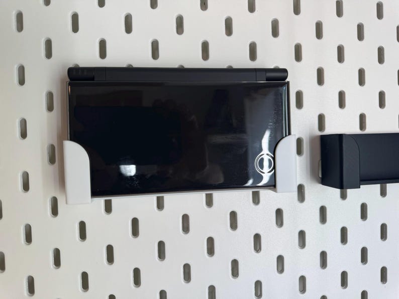 May include: A black handheld gaming console is mounted on a white pegboard using a white bracket. The console is closed and has a reflective surface. A second black bracket is also mounted on the pegboard.