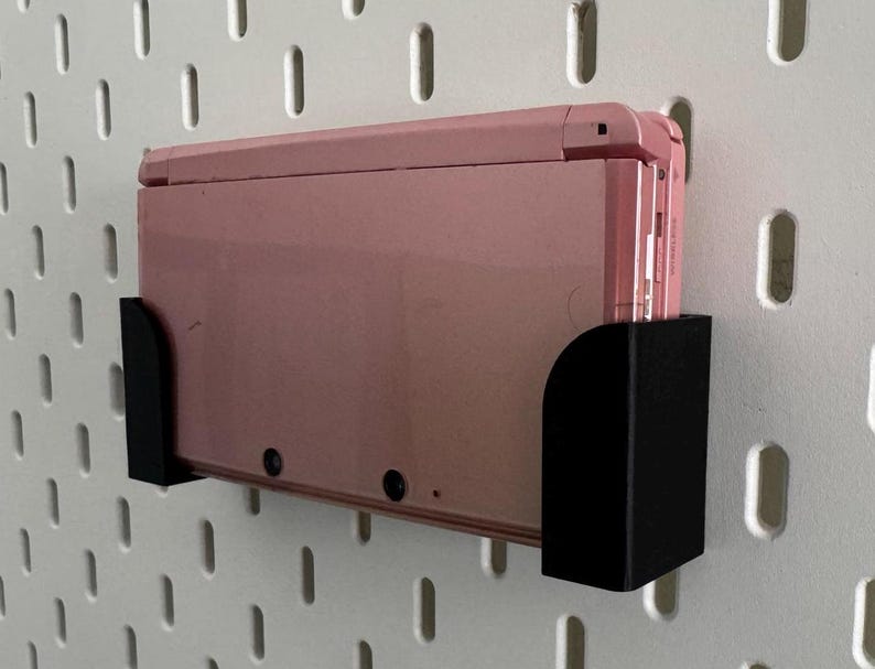 Pode incluir: Um console Nintendo Switch rosa est&aacute; montado em um suporte de parede de pegboard preto. O suporte tem um design simples e &eacute; feito de pl&aacute;stico resistente.