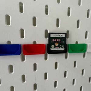 May include: Four small plastic storage bins in blue, red, green, and white are mounted on a white pegboard. A Nintendo DS game cartridge is mounted in the centre of the pegboard. The game cartridge is for the game 42 All-Time Classics.