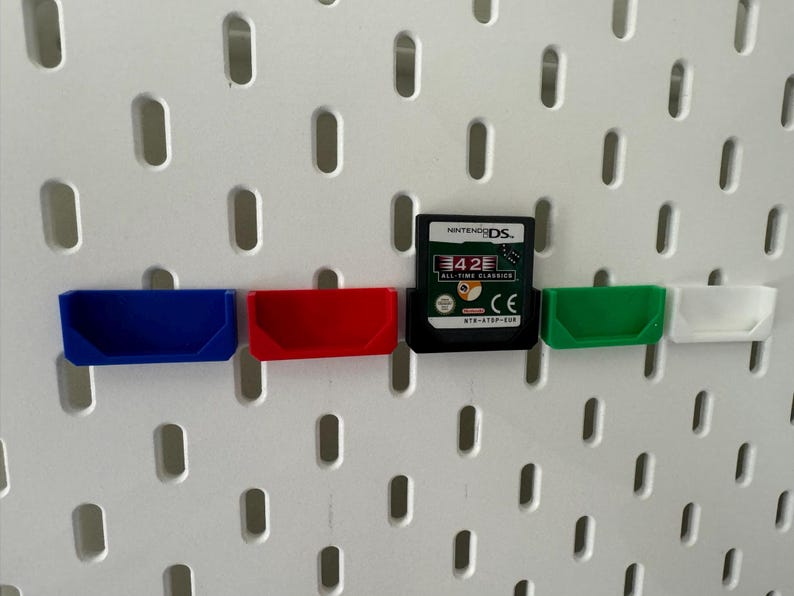 May include: Four small plastic storage bins in blue, red, green, and white are mounted on a white pegboard. A Nintendo DS game cartridge is mounted in the centre of the pegboard. The game cartridge is for the game 42 All-Time Classics.
