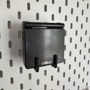May include: A black Nintendo Game Boy Advance SP handheld console mounted on a white pegboard. The console is held in place by a black bracket. The Nintendo logo is visible on the console.