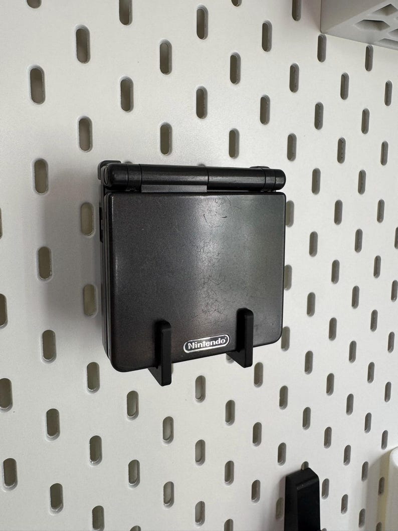 May include: A black Nintendo Game Boy Advance SP handheld console mounted on a white pegboard. The console is held in place by a black bracket. The Nintendo logo is visible on the console.