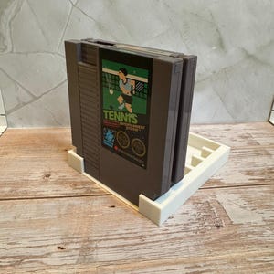 May include: A gray Nintendo Entertainment System (NES) game cartridge for the game "Tennis" is displayed in a white holder. The game cartridge has a green and black label with a tennis player graphic. The holder is on a wooden surface.