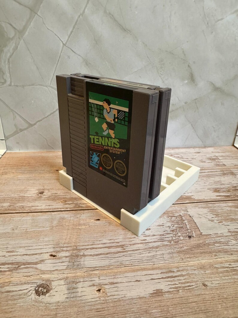 May include: A gray Nintendo Entertainment System (NES) game cartridge for the game "Tennis" is displayed in a white holder. The game cartridge has a green and black label with a tennis player graphic. The holder is on a wooden surface.