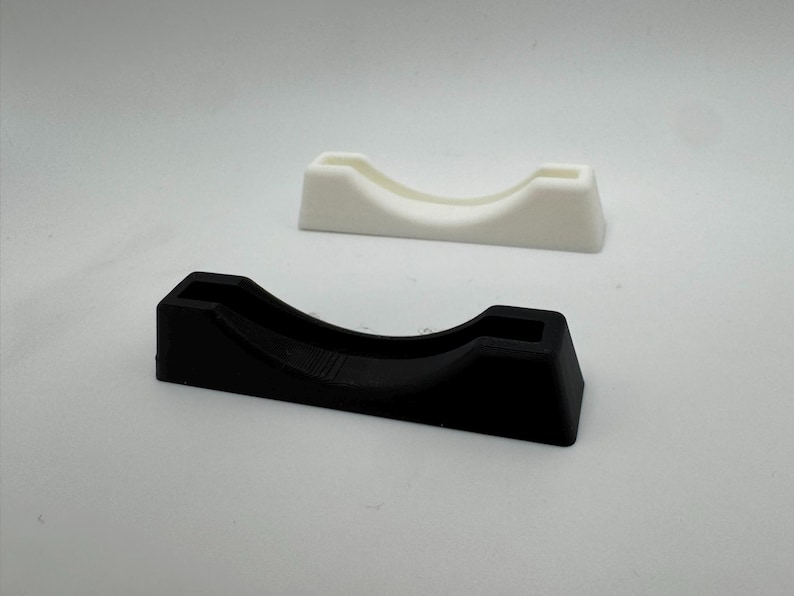 Puede incluir: Dos peque&ntilde;os expositores curvos. Uno es negro, el otro blanco. Ambos tienen una base rectangular y una parte superior curva con una peque&ntilde;a abertura cuadrada. Los expositores est&aacute;n dise&ntilde;ados para sostener objetos peque&ntilde;os.
