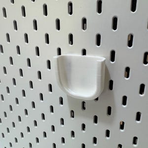 K&ouml;nnte beinhalten: Ein wei&szlig;er, 3D-gedruckter Becherhalter f&uuml;r eine Lochwand. Der Halter ist an einer wei&szlig;en Lochwand mit gleichm&auml;&szlig;ig verteilten, oval geformten L&ouml;chern befestigt. Der Becherhalter hat ein gebogenes, U-f&ouml;rmiges Design.