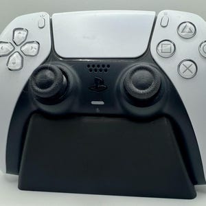 May include: A white PlayStation 5 DualSense controller with black analogue sticks and face buttons, resting on a black charging stand.