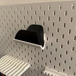 May include: A black charging dock with a white base is mounted on a white pegboard. The pegboard has evenly spaced oval holes. A white shelf with a wavy design is below the dock. The items are for home organization.