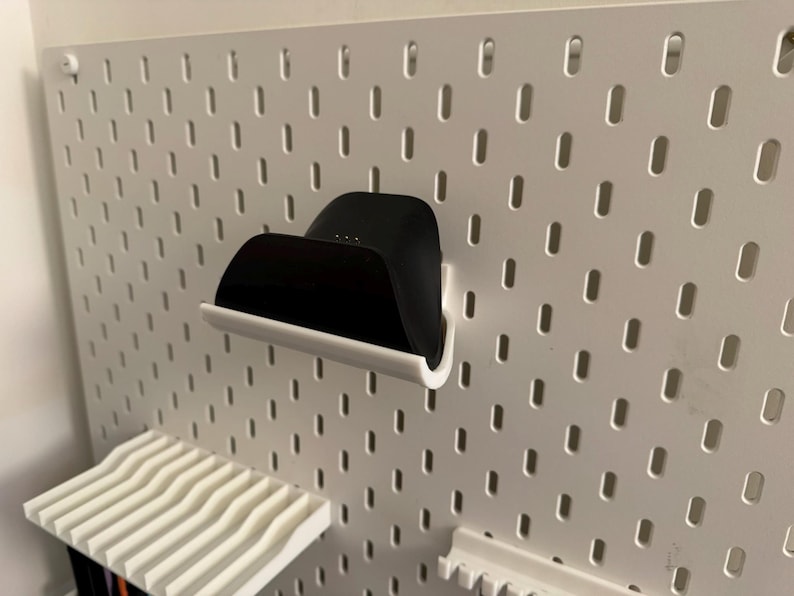 May include: A black charging dock with a white base is mounted on a white pegboard. The pegboard has evenly spaced oval holes. A white shelf with a wavy design is below the dock. The items are for home organization.