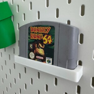 May include: A gray Nintendo 64 video game cartridge for Donkey Kong 64, displayed on a white shelf. The cartridge features the game's logo with a picture of Donkey Kong. The background is a white pegboard.