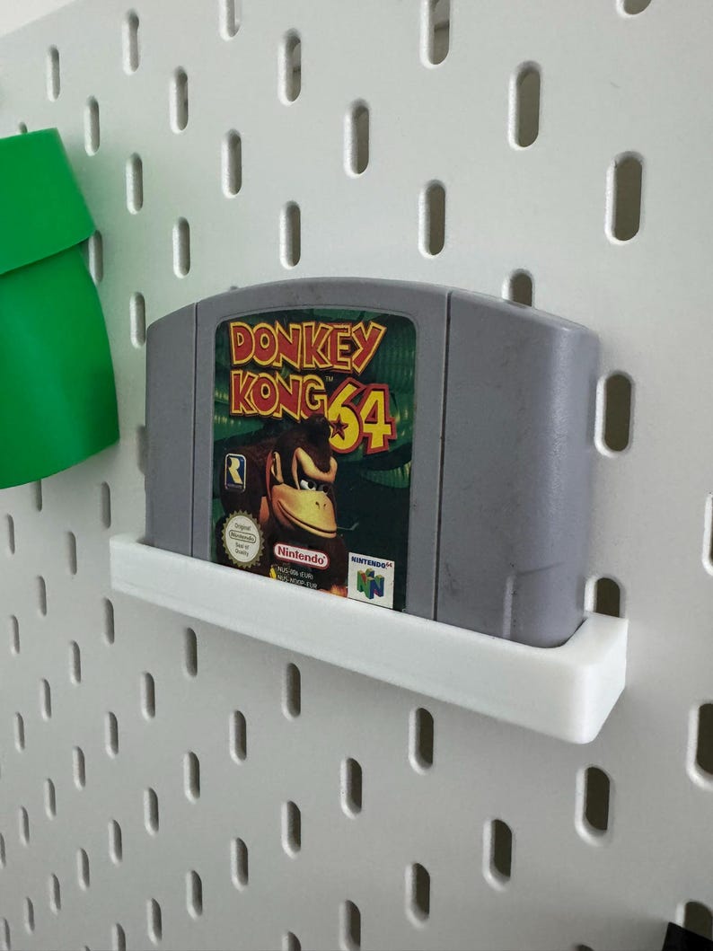 May include: A gray Nintendo 64 video game cartridge for Donkey Kong 64, displayed on a white shelf. The cartridge features the game's logo with a picture of Donkey Kong. The background is a white pegboard.