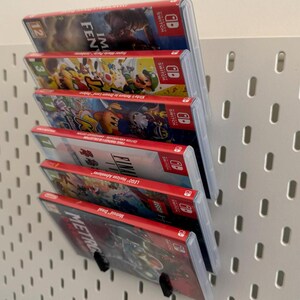 Pu&ograve; includere: Una pila di sei cartucce di gioco per Nintendo Switch, esposte su una parete di bacheca bianca. Le cartucce sono tutte rosse e nere, con vari titoli di gioco stampati su di esse. I titoli includono "Lego Horizon Adventures", "Final Fantasy", "Kirby Star Allies", "Mario Kart 8 Deluxe", "Super Mario Odyssey" e "I am Setsuna".