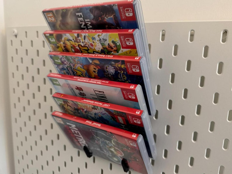 Pu&ograve; includere: Una pila di sei cartucce di gioco per Nintendo Switch, esposte su una parete di bacheca bianca. Le cartucce sono tutte rosse e nere, con vari titoli di gioco stampati su di esse. I titoli includono "Lego Horizon Adventures", "Final Fantasy", "Kirby Star Allies", "Mario Kart 8 Deluxe", "Super Mario Odyssey" e "I am Setsuna".