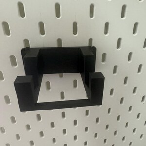 K&ouml;nnte beinhalten: Schwarzer Wandhaken aus Kunststoff mit quadratischer Form. Der Haken ist an einer wei&szlig;en Lochwand befestigt.
