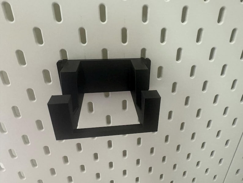 K&ouml;nnte beinhalten: Schwarzer Wandhaken aus Kunststoff mit quadratischer Form. Der Haken ist an einer wei&szlig;en Lochwand befestigt.