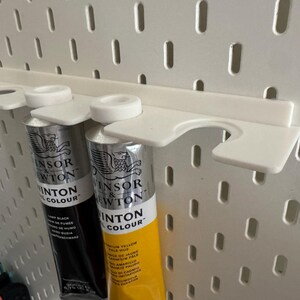 May include: A white plastic paint tube holder attached to a pegboard. Two tubes of Winsor & Newton paint are held in place. One tube is black, and the other is yellow. The pegboard has evenly spaced holes.