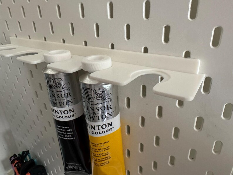 May include: A white plastic paint tube holder attached to a pegboard. Two tubes of Winsor & Newton paint are held in place. One tube is black, and the other is yellow. The pegboard has evenly spaced holes.