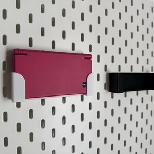 K&ouml;nnte beinhalten: Eine magenta Nintendo DS-Handheld-Spielkonsole ist mit einem wei&szlig;en Halter an einer wei&szlig;en Lochwand befestigt. Rechts ist ein schwarzer Halter montiert. Die Lochwand hat ein Muster aus gleichm&auml;&szlig;ig verteilten ovalen L&ouml;chern.
