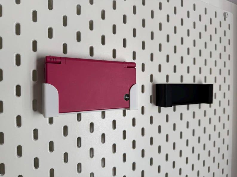K&ouml;nnte beinhalten: Eine magenta Nintendo DS-Handheld-Spielkonsole ist mit einem wei&szlig;en Halter an einer wei&szlig;en Lochwand befestigt. Rechts ist ein schwarzer Halter montiert. Die Lochwand hat ein Muster aus gleichm&auml;&szlig;ig verteilten ovalen L&ouml;chern.