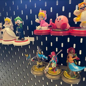 May include: A collection of colorful Nintendo character figurines displayed on a black pegboard wall. The figurines include Mario, Luigi, Peach, Toad, Link, Zelda, and Kirby.