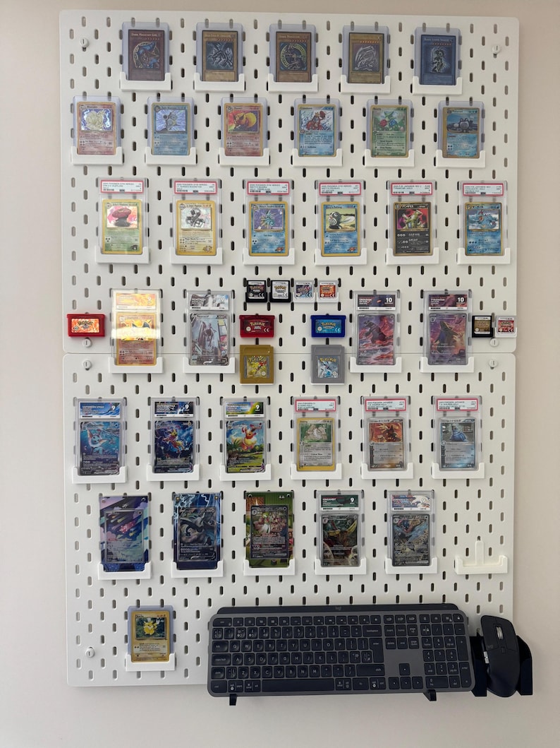 K&ouml;nnte beinhalten: Eine wei&szlig;e Lochwand mit einer Vielzahl von Pok&eacute;mon-Karten, die in sch&uuml;tzenden Plastikh&uuml;llen ausgestellt sind. Die Karten sind in Reihen und Spalten angeordnet, wobei einige Karten holografische Designs aufweisen. Eine Tastatur und eine Maus sind ebenfalls in der unteren rechten Ecke des Bildes zu sehen.