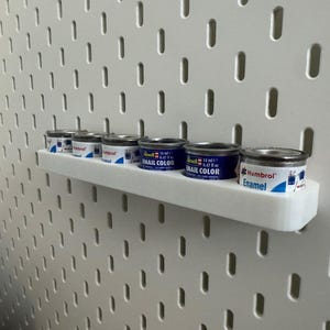 May include: A white pegboard with a white shelf holding several small paint cans. The cans are silver with blue and white labels. The text "Enamel Color" and "Humbrol" are visible on the cans.