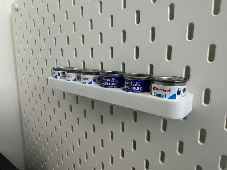 May include: A white pegboard with a white shelf holding several small paint cans. The cans are silver with blue and white labels. The text "Enamel Color" and "Humbrol" are visible on the cans.