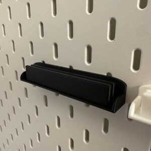 Pode incluir: Uma prateleira preta para painel perfurado &eacute; montada em um painel perfurado branco. A prateleira &eacute; retangular com uma frente curva e foi projetada para guardar itens pequenos. O painel perfurado tem orif&iacute;cios ovais espa&ccedil;ados.